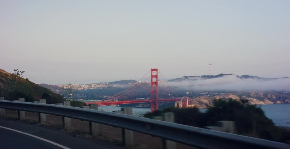 A classic car leads the way on a winding coastal road, revealing a spectacular view of the Golden Gate Bridge and a foggy San Francisco skyline