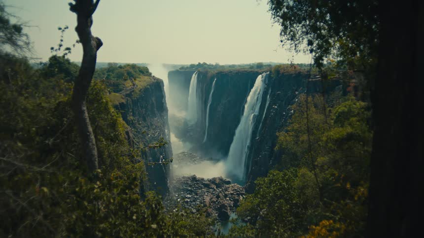 A wide, static shot of the magnificent Victoria Falls cascading into a deep, rocky gorge, viewed from behind jungle foliage