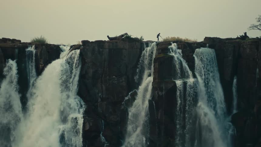 A lone fisherman casts a line from the edge of a vast, powerful waterfall cascading over a sheer rock cliff