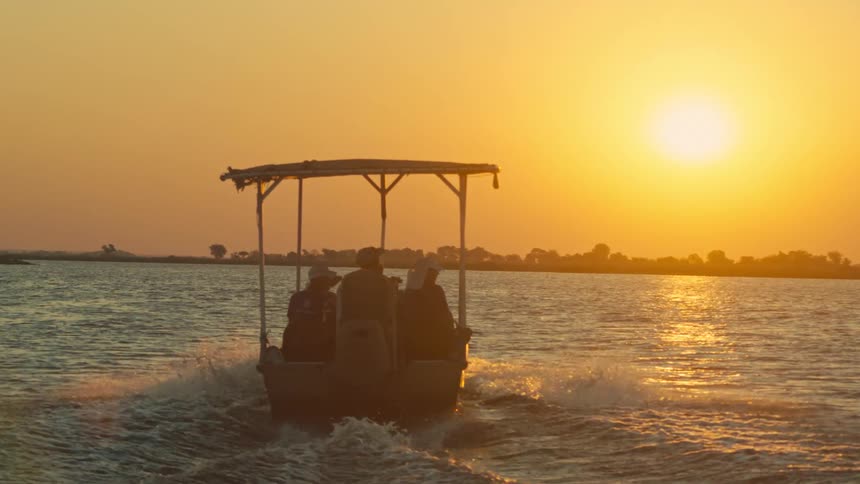 A small tour boat with silhouetted passengers glides across a wide river into the beautiful golden sunset
