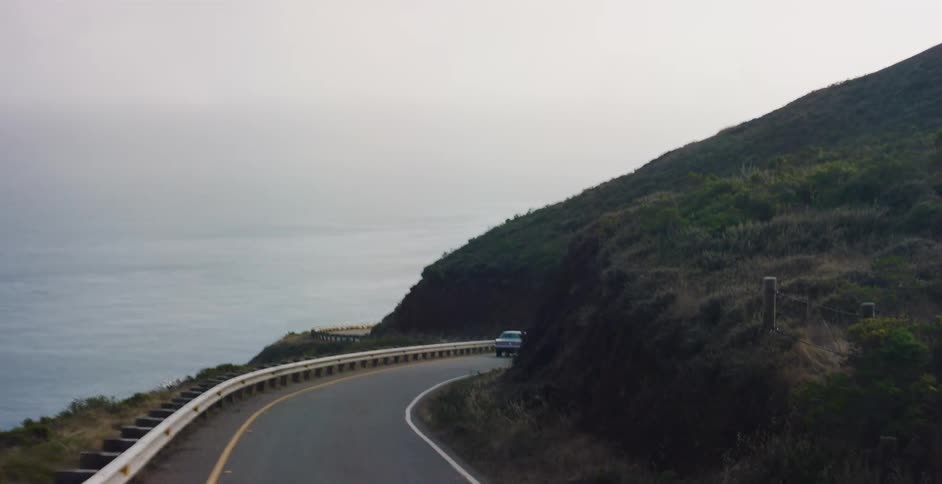 A vintage car navigates a winding coastal highway with scenic views of the ocean on a hazy day