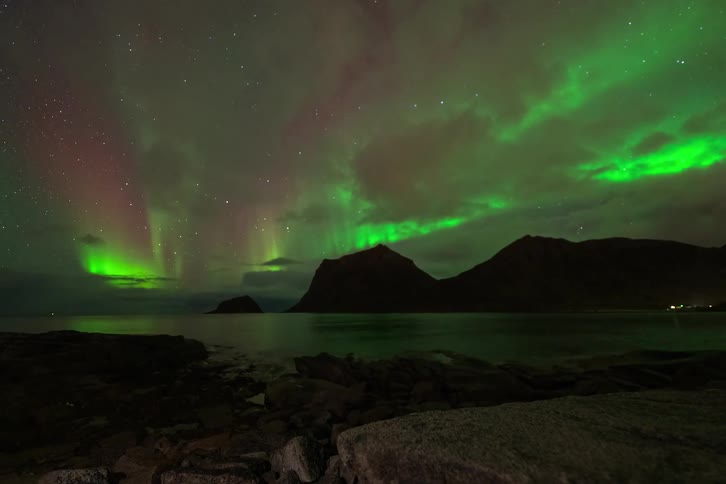 A stunning timelapse of the green and magenta Aurora Borealis dancing across a starry night sky over silhouetted mountains and a tranquil sea