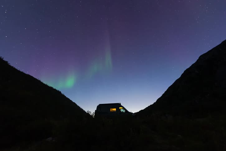 A person celebrates on top of a camper van as the vibrant Northern Lights dance across the starry night sky between two mountains
