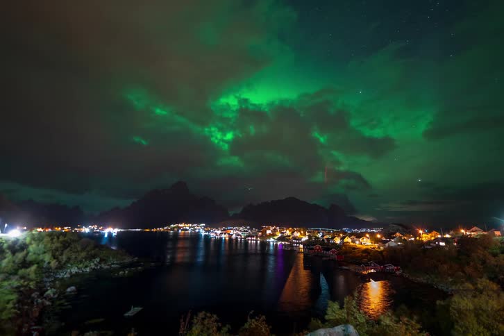 A stunning timelapse shows the vibrant green aurora borealis dancing in the starry night sky above an illuminated coastal town nestled by mountains and a tranquil bay