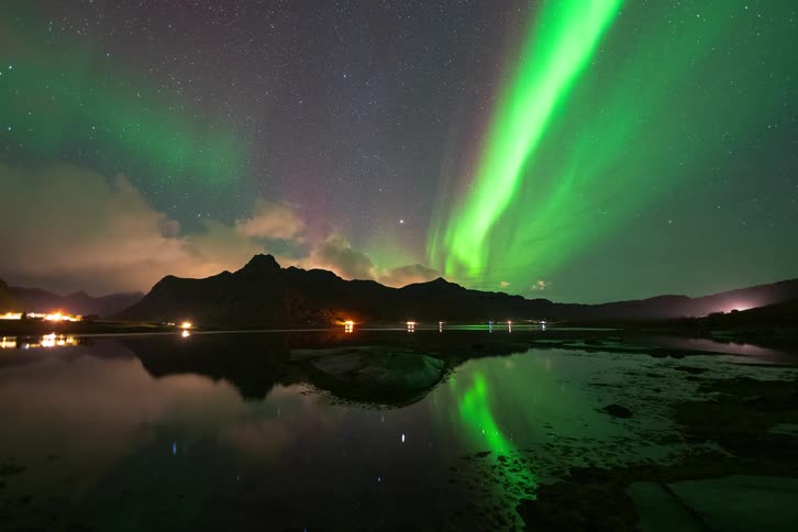 A stunning timelapse captures the vibrant green Aurora Borealis dancing across the starry night sky over a mountain range, reflecting in the still water below