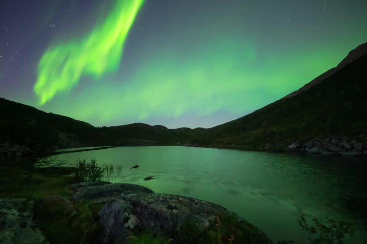 A stunning time-lapse captures the vibrant green Northern Lights dancing in the starry night sky, reflecting beautifully over a tranquil mountain lake