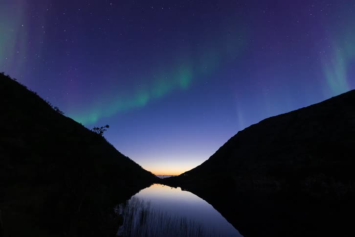 A stunning timelapse captures the vibrant green and purple Aurora Borealis dancing across the starry night sky, perfectly reflected in a calm lake