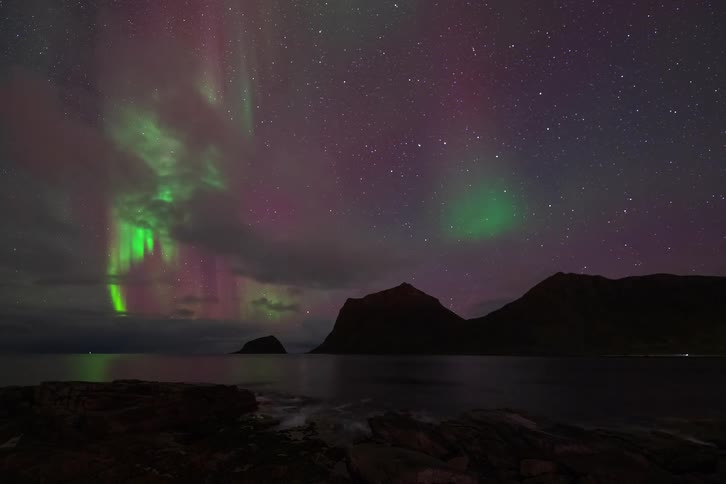 A time-lapse captures the vibrant green and purple Aurora Borealis dancing in the starry night sky over a calm sea and silhouetted mountains