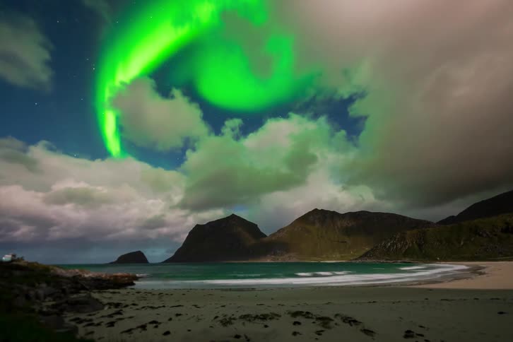 A stunning night timelapse shows the vibrant green Aurora Borealis dancing through fast-moving clouds over a tranquil beach and mountains