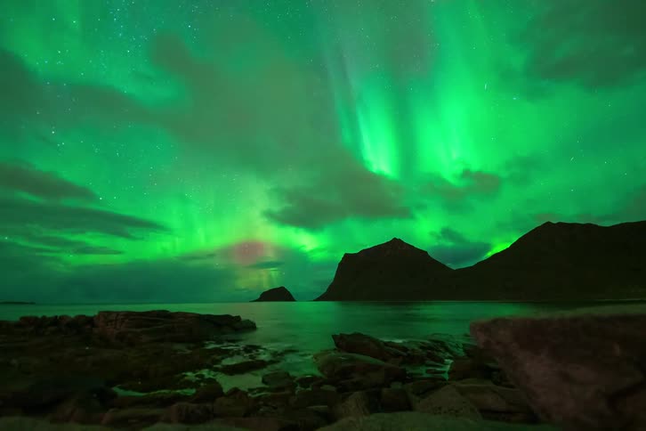 A breathtaking time-lapse captures the vibrant green Aurora Borealis dancing in the night sky over a serene, rocky coastline and distant mountains