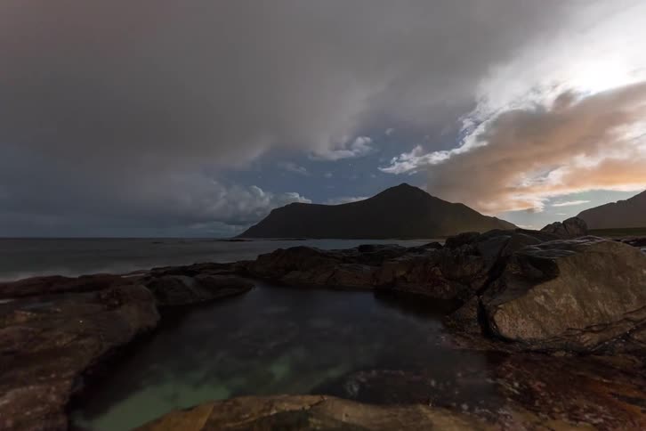 A stunning night time-lapse captures the Aurora Borealis and stars above a rugged, rocky coastline as dramatic clouds roll across the sky