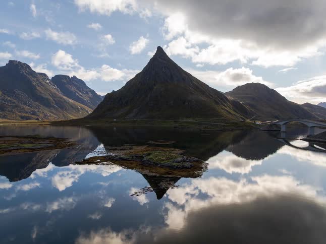 An aerial view pulls back to reveal a sharp mountain peak perfectly mirrored in the still waters of a fjord in the Lofoten Islands