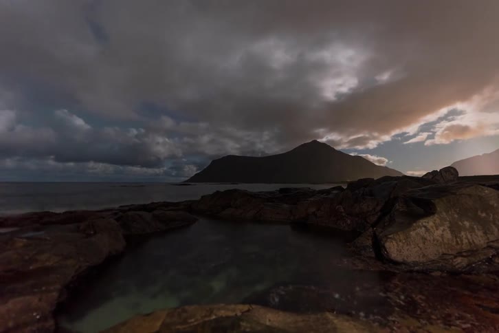 A breathtaking time-lapse shows the moon setting and the aurora borealis emerging from behind moving clouds over a rocky coast, with the night sky reflected in a tidal pool