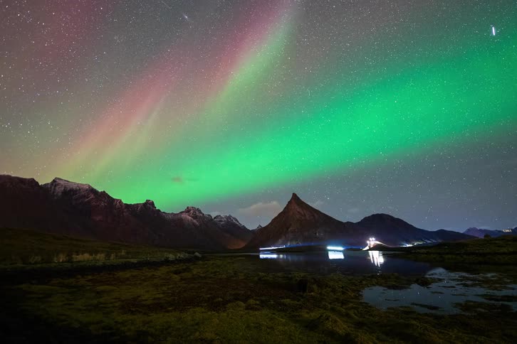 A vibrant timelapse of the green and pink Aurora Borealis dancing across a starry night sky above a majestic mountain range and its reflection in a lake