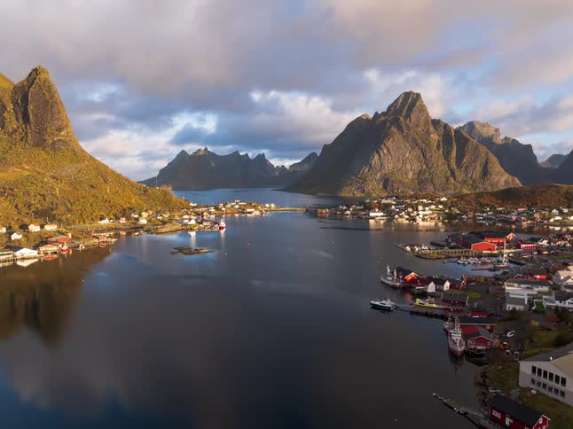 A breathtaking aerial shot pulls back to reveal the iconic Norwegian fishing village of Reine, perfectly reflected in the calm fjord amidst towering mountains during a golden sunrise