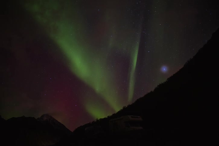 A vibrant time-lapse shows the green Aurora Borealis dancing in the starry night sky above a camper van in the mountains