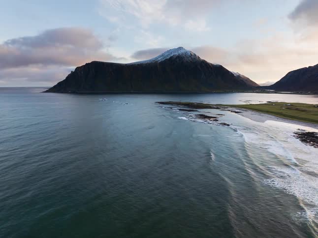 An aerial view pulls back over a stunning Norwegian coastline, revealing gentle ocean waves and a majestic snow-capped mountain