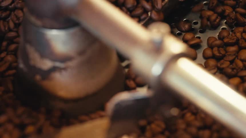 A close-up slow-motion view shows rich, brown coffee beans tumbling in an industrial roasting machine