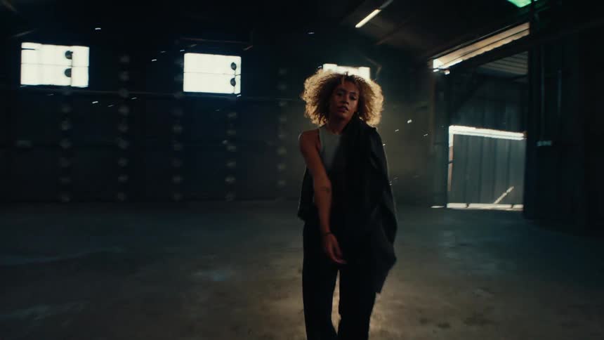 A confident woman with curly hair walks towards the camera and throws her jacket in a sunlit, industrial warehouse