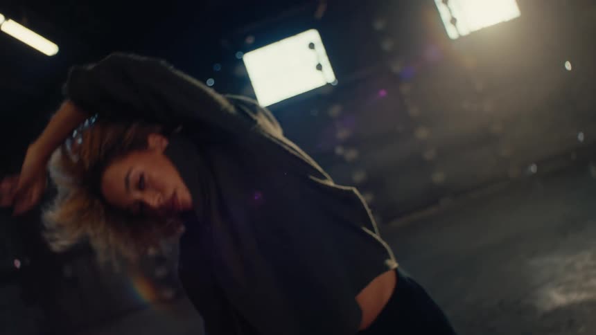Low-angle slow motion of a woman with curly hair dancing freely in a large, empty warehouse with dramatic lighting
