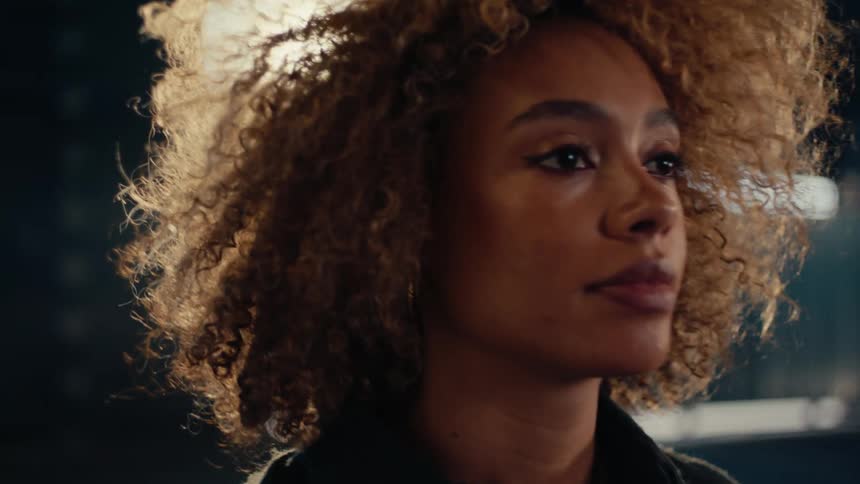 A striking woman with curly hair and eyeliner comes into focus with a serious expression in a dimly lit, atmospheric setting