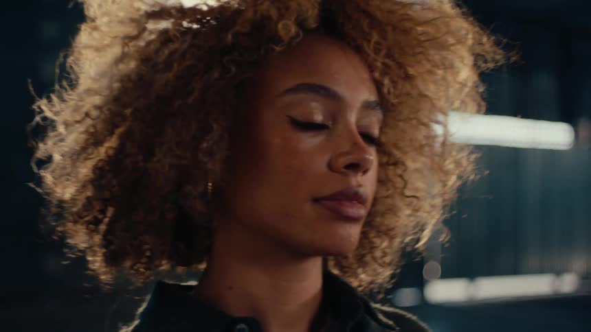 A slow-motion, backlit close-up reveals a stylish woman with curly blonde hair looking pensively into the distance