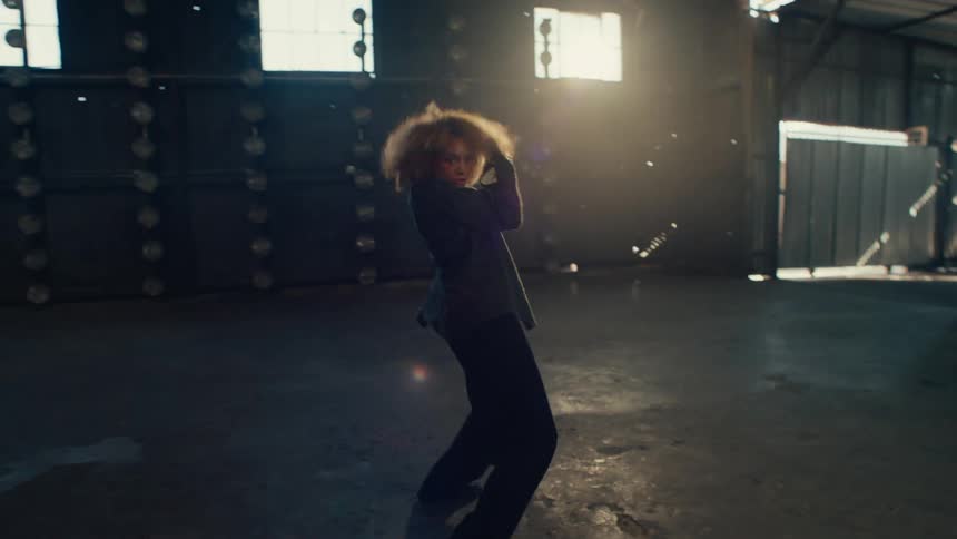 A passionate female dancer with an afro performs a dynamic and expressive freestyle dance in a large, dark warehouse in slow motion