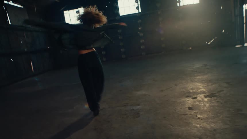 Slow-motion footage of a talented female dancer performing an interpretive dance in a vast, dark, backlit warehouse