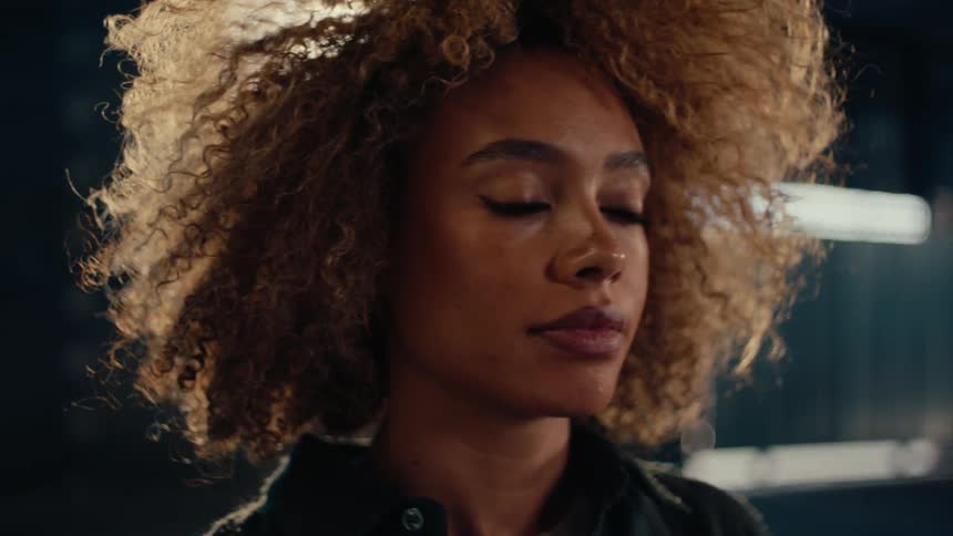Cinematic close-up of a stylish woman with curly blonde hair looking thoughtful in a dark, backlit room