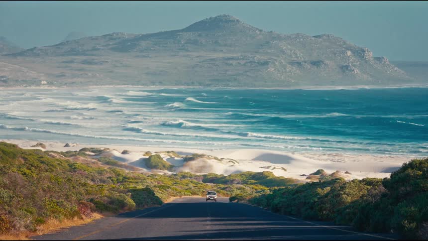 A single vehicle drives up a coastal road, revealing a beautiful vista of a sandy beach, turquoise ocean, and a distant mountain