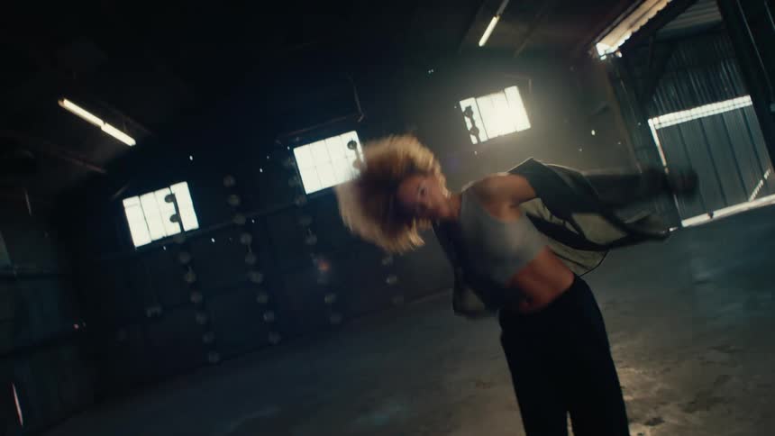 Slow motion of a passionate female dancer with an afro performing an energetic freestyle in a dark, backlit warehouse