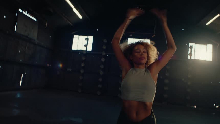 Slow motion shot of a stylish woman with curly hair dancing passionately in an empty, backlit industrial warehouse