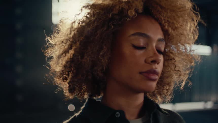 A beautiful woman with curly hair dramatically tosses her head back in slow motion in a moody, backlit industrial space