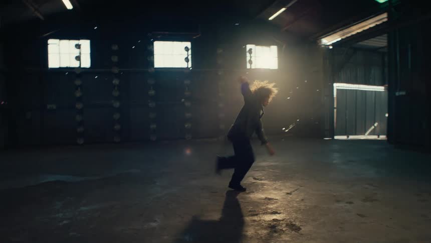 A stylish woman with afro hair performs an energetic solo dance in a large, empty, backlit warehouse with stage lights