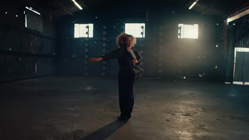 Expressive female dancer with an afro dances solo in a large, empty industrial warehouse in slow motion