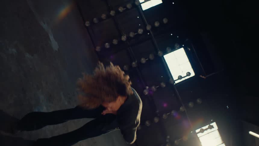 A vertical slow-motion shot shows a stylish woman with an afro dancing gracefully as she floats in a dark, sunlit warehouse