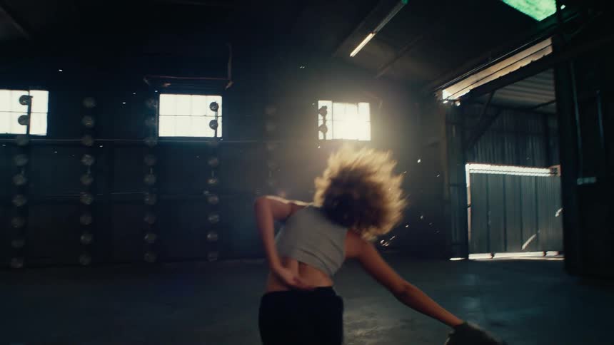 A stylish young woman with an afro expressively dances and removes her jacket in slow motion inside a large, empty warehouse with dramatic backlighting
