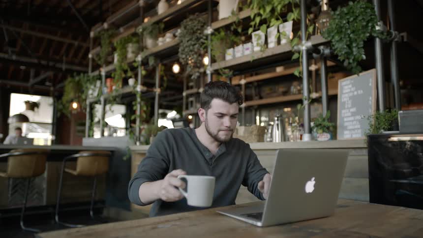 A man is drinking from a cup and working on a laptop in a coffee shop