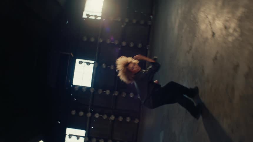 A rotating camera captures a woman with curly hair gracefully moving on the floor of a dark warehouse, creating a mesmerizing anti-gravity illusion