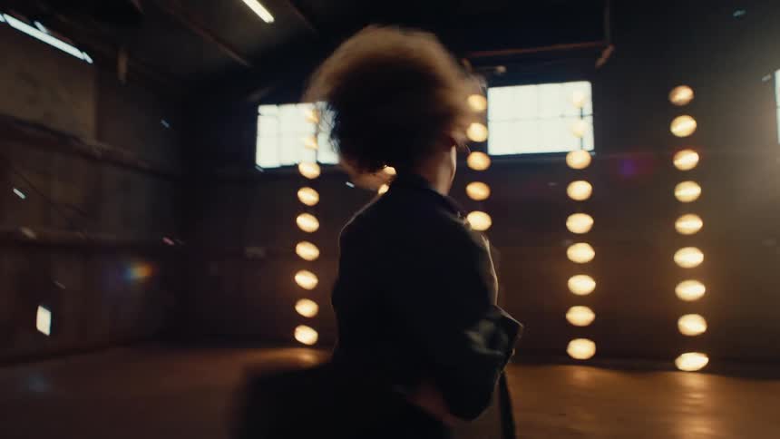 A talented female dancer with an afro performs a passionate and emotional contemporary dance in a large, dark, backlit warehouse