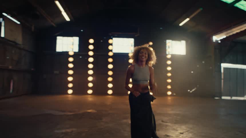 A happy, stylish woman with curly hair walks into an empty industrial warehouse with rows of bright lights