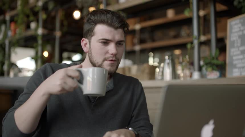 A man drinks coffee while working on his laptop in a coffee shop