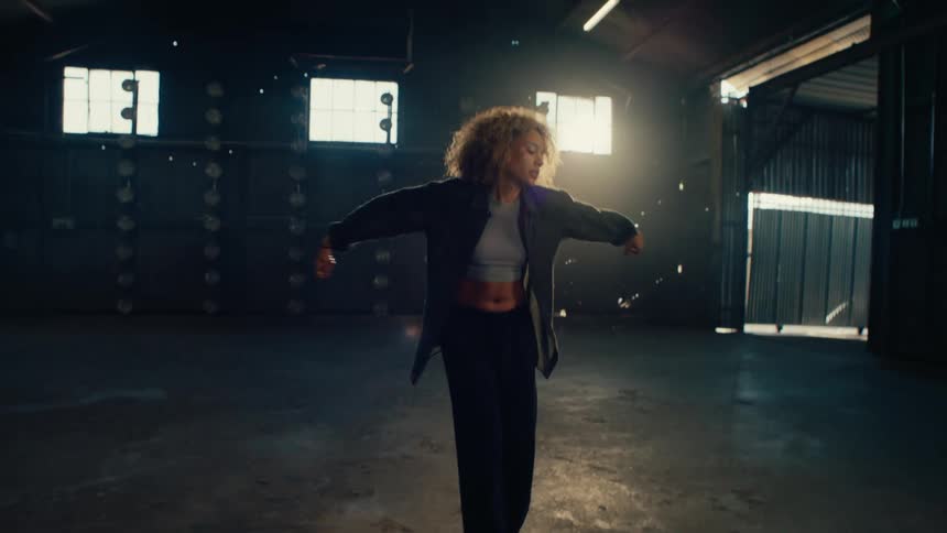 A stylish woman with an afro performs an expressive modern dance routine in slow motion inside a large, dark, backlit warehouse