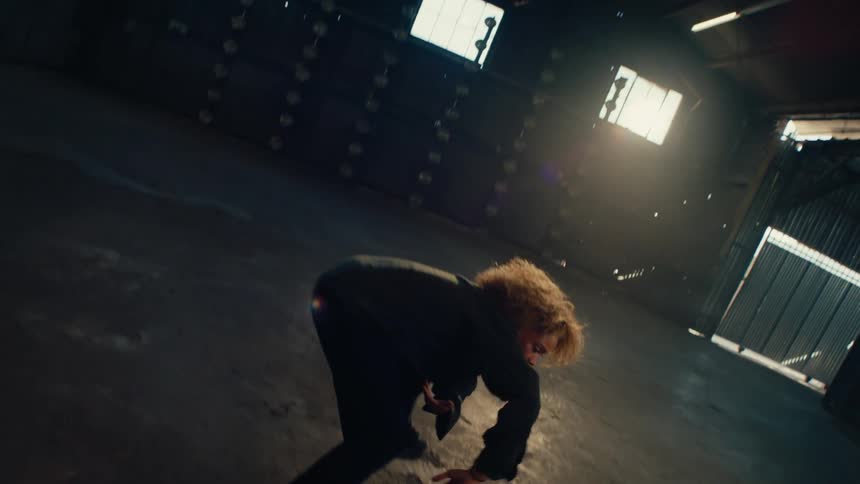 An expressive female dancer with curly hair performs a contemporary routine in a dark, sunlit warehouse