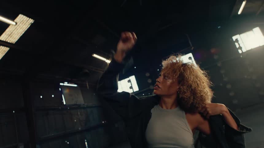 Slow motion shot of a woman with curly hair putting on a jacket and beginning to dance in a moody, backlit industrial warehouse