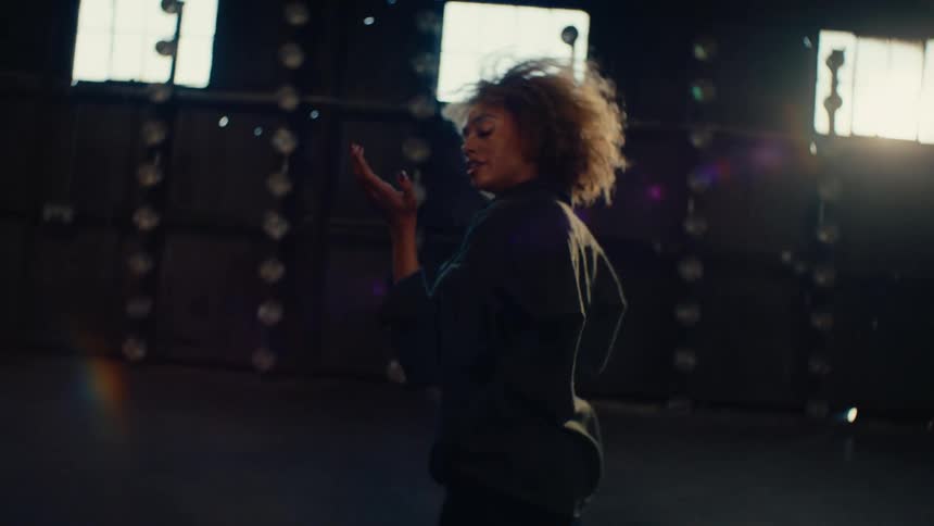 A stylish woman with an afro dances in slow motion inside a dark, empty warehouse as the camera spins around her