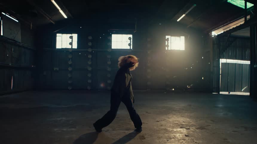 A female dancer walks towards the camera in a dusty, backlit warehouse and begins an energetic, expressive contemporary dance routine