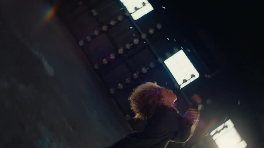 A surreal, slow-motion shot of a dancer with an afro defying gravity as they float down from the ceiling of a vast, sunlit industrial warehouse