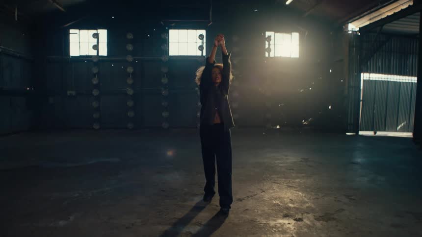 A stylish dancer with curly hair performs expressive, fluid movements in slow motion inside a vast, sunlit industrial warehouse