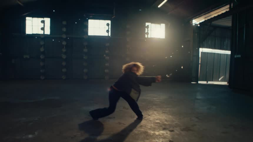 A talented female dancer with curly hair performs an emotional contemporary routine in a vast, empty warehouse backlit by dramatic sunlight